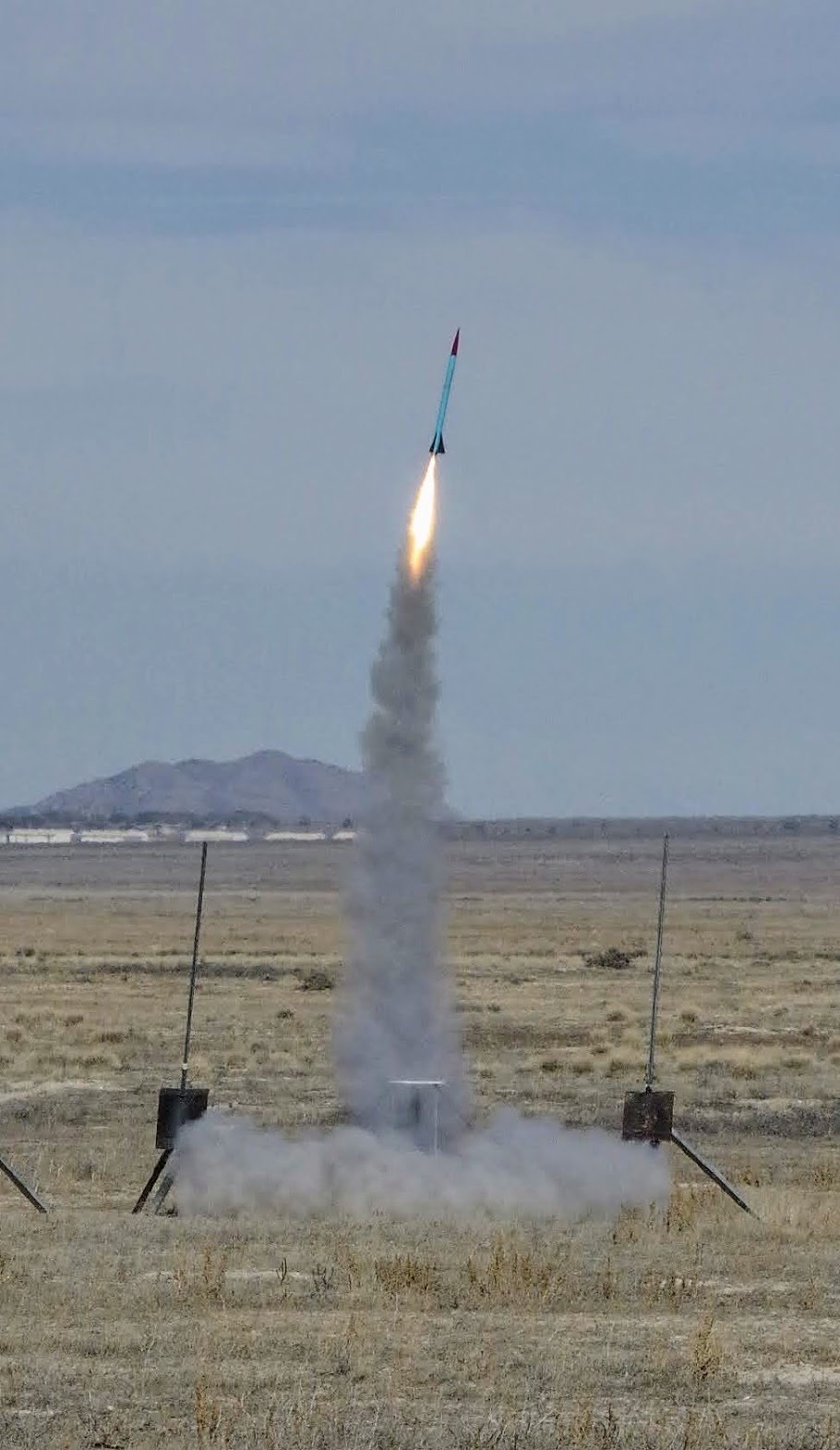 The teal and red airframe climbing away above a tall column of smoke, launch towers small below it.